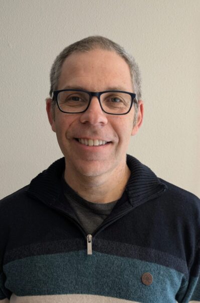 Headshot of a person wearing glasses and a striped sweater, smiling in front of a plain background.